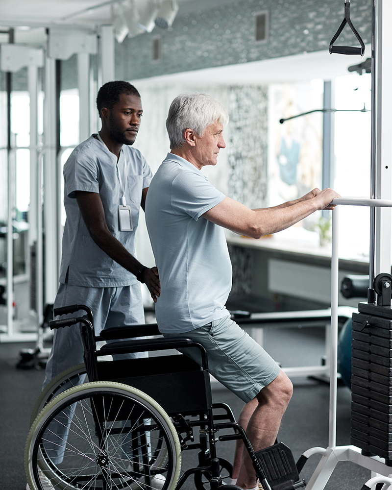 Physical therapist assisting an older man in a wheelchair with leg exercises using gym equipment.
